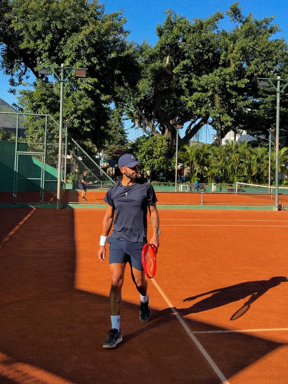 João caminhando em uma quadra de saibro, de raquete na mão